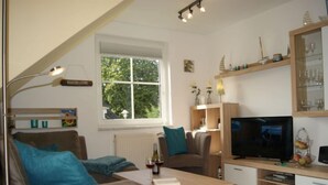 Ein modernes Wohnzimmer mit Sofa, Couchtisch, Fernseher und einem Fenster mit Blick auf Grünflächen.
