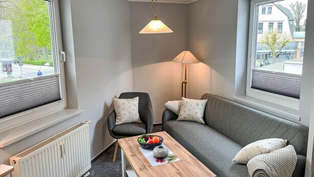 Ein modernes, kompaktes Wohnzimmer mit grauem Sofa, einem Holztisch mit Obstschale, einer Stehlampe und einer Heizung.
