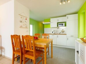 Cozinha moderna com armários brancos e uma parede verde de destaque, uma mesa de jantar de madeira com cadeiras e um micro-ondas acima dos armários.