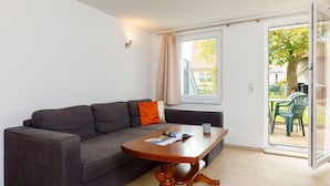 Een woonkamer met een grijze bank, een houten salontafel en een schuifdeur naar een buitenruimte met stoelen en een boom.