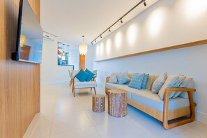 Una sala moderna con un sofá, dos taburetes de madera, un televisor montado en una pared de madera y un comedor al fondo.