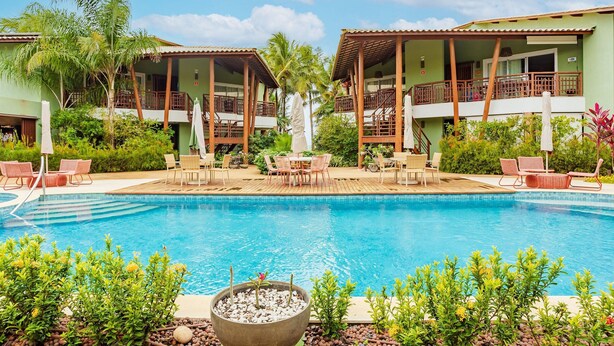 Zona de piscina con sillones reclinables y una pequeña mesa, rodeada de vegetación y edificios de dos pisos con balcones.