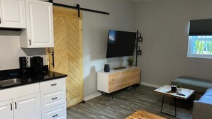 Eine moderne Küche mit weißen Schränken, einer schwarzen Arbeitsplatte und einem Holzboden im Fischgrätmuster. Im Wohnbereich befindet sich ein Flachbildfernseher an der Wand, ein kleiner Holztisch und ein graues Sofa.