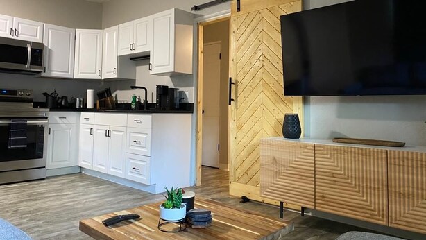 Eine moderne Küche mit weißen Schränken, einer Mikrowelle und einem Herd. Eine Holztür mit Fischgrätmuster führt in einen anderen Raum. Ein Flachbildfernseher ist über einem Holzschrank an der Wand befestigt.
