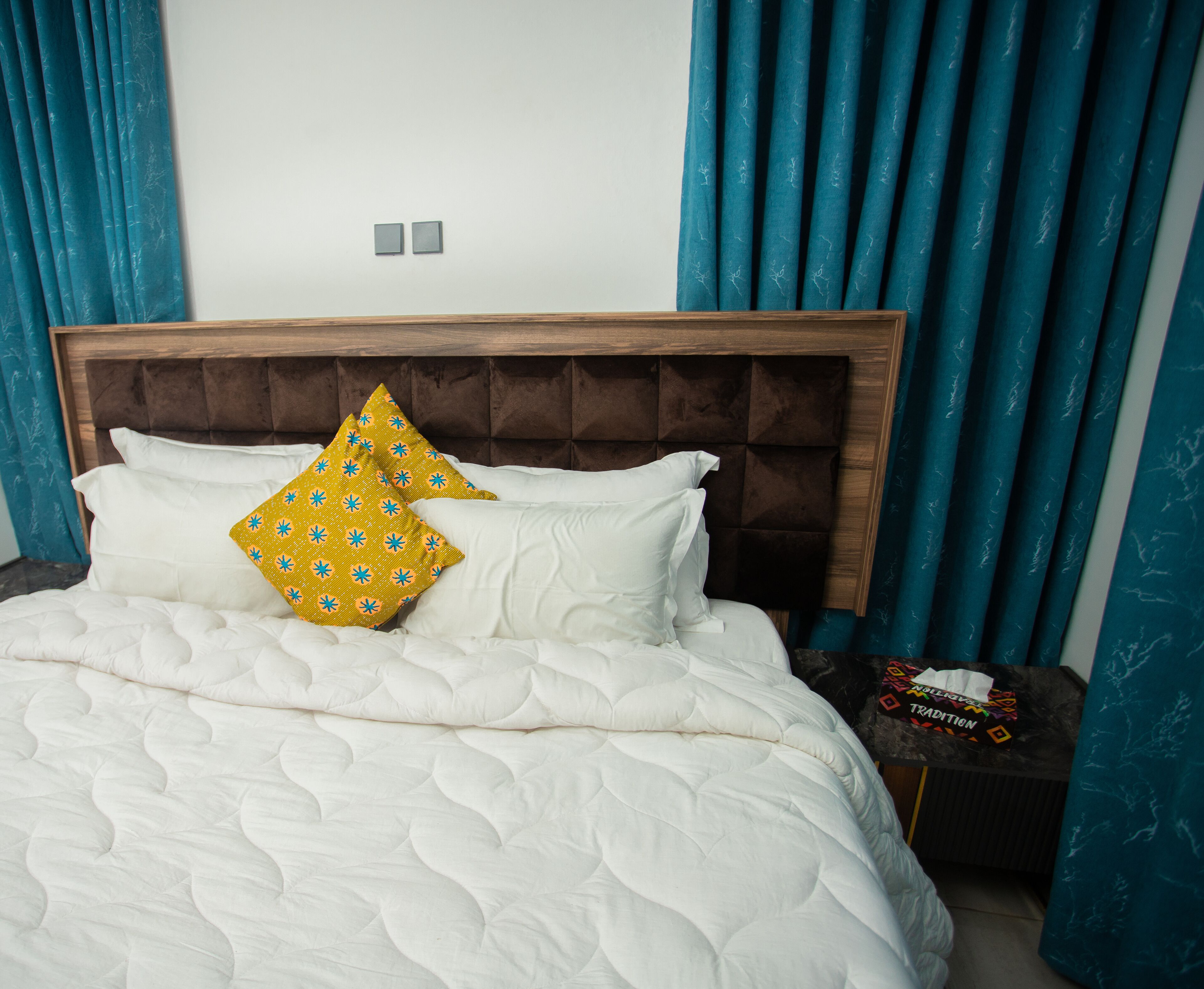 A bed with white bedding and two pillows, one yellow and one white, in a room with teal curtains and a wooden headboard.