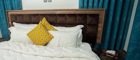 A bed with white bedding and two pillows, one yellow and one white, in a room with teal curtains and a wooden headboard.