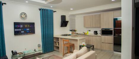 A modern living room with a sofa, a TV mounted on the wall, and a kitchen area with wooden cabinets and a stove.