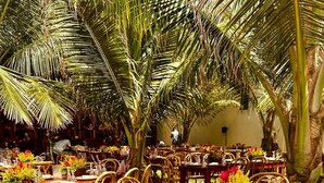 An outdoor dining area with wooden tables set for multiple guests, surrounded by palm trees. - Reddington Beach Resort (Anloga)