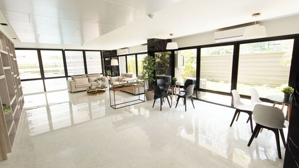 Amplia sala de estar con diseño moderno, que incluye un conjunto de sofá, una mesa de centro y una mesa de comedor con sillas. Grandes ventanales ofrecen abundante luz natural y vistas al exterior.