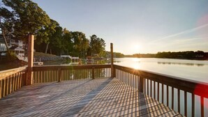 A wooden deck with a railing overlooking a calm lake, surrounded by trees and buildings. - River District Retreat—Walk to Downtown Belmont! (Belmont)