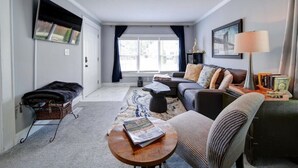 A living room with a sofa, armchair, coffee table, and a small table with a lamp. There is a window with curtains, a fireplace, and a bookshelf. - River District Retreat—Walk to Downtown Belmont! (Belmont)
