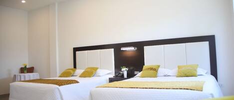 Una habitación de hotel con dos camas, cada una con ropa de cama blanca y almohadas amarillas. Hay una mesita de noche con una lámpara y una planta pequeña. La habitación tiene un diseño moderno, con un cabecero oscuro e iluminación empotrada en el techo.