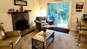 A living room with a fireplace, a sofa, a coffee table, and a large sliding glass door leading to a covered outdoor area. - Peaceful Family Retreat – Spacious 4BR Home - 5 acres of forest (Inlet)