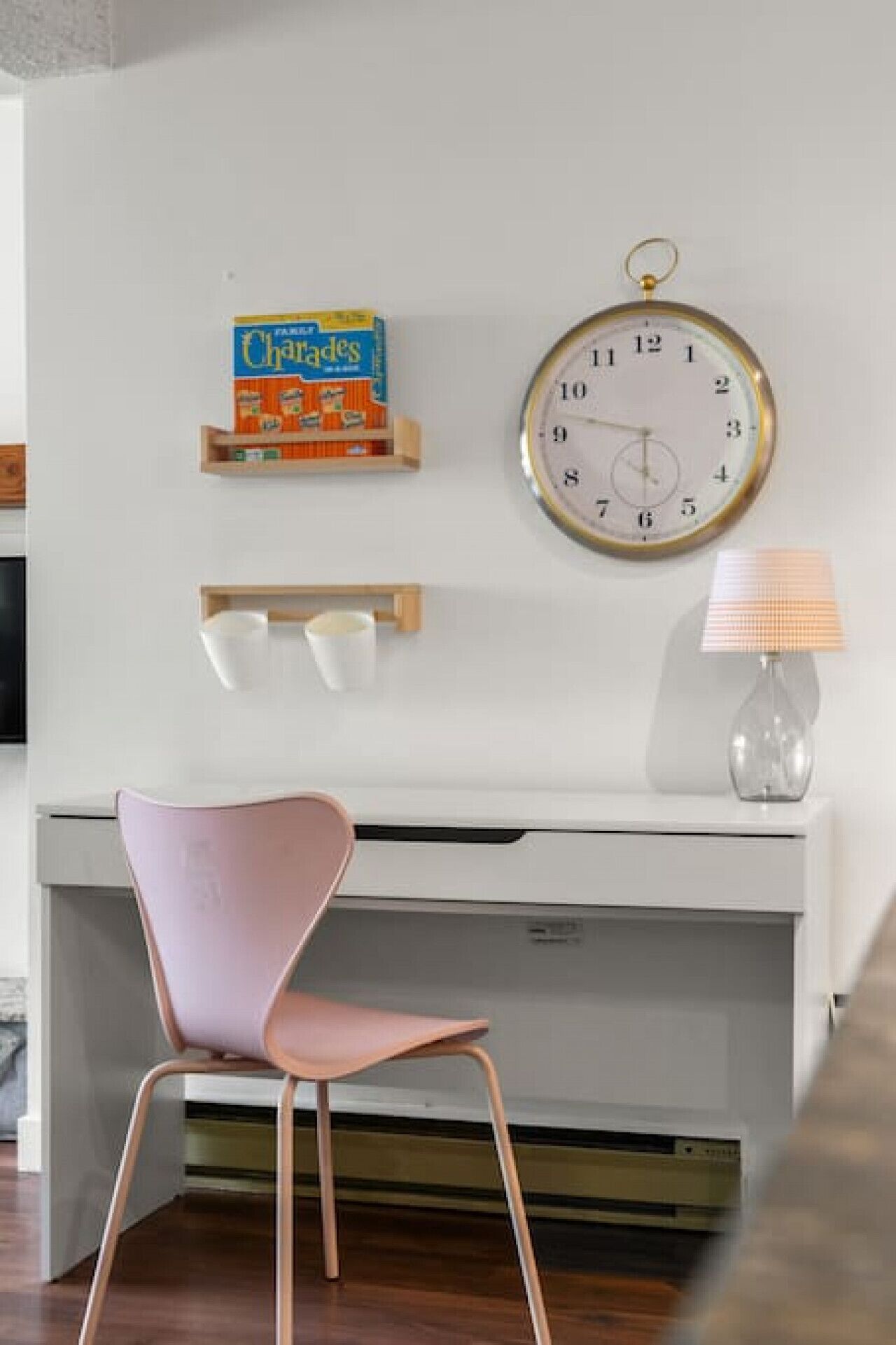 Une pièce moderne avec un bureau blanc, une chaise rose, une horloge ronde accrochée au mur, une lampe et une boîte de céréales posée sur le mur.