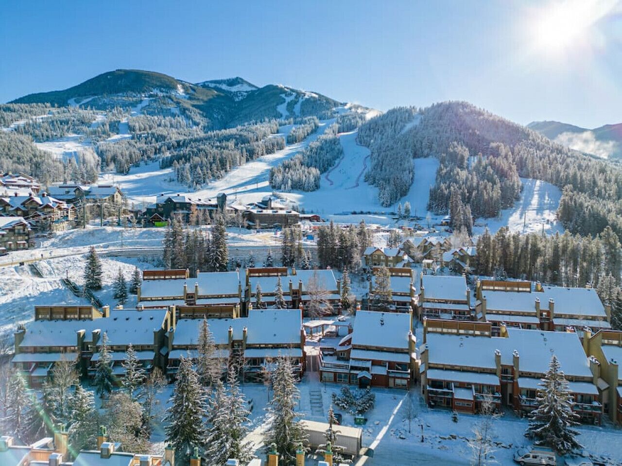 A snow-covered resort with multiple chalet-style buildings, a ski slope in the background, and a mountainous landscape.