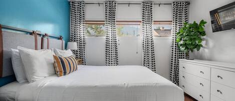 A modern bedroom with a large bed, white bedding, a blue accent wall, a white dresser, and a window with patterned curtains.