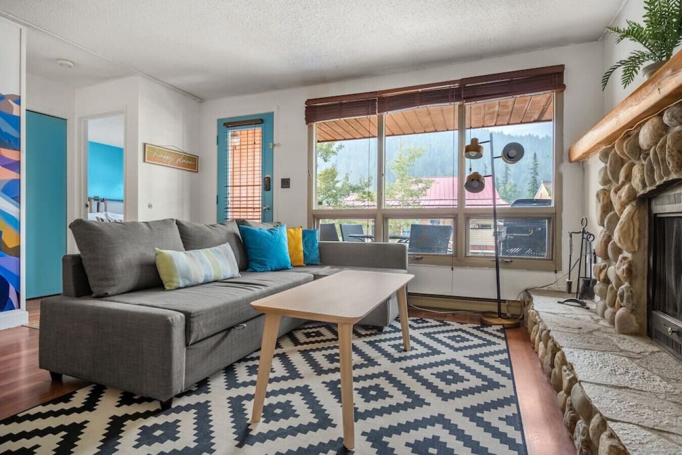 A cozy living room with a gray sofa, a wooden coffee table, and a patterned rug. There's a stone fireplace and a large window with a view of the outside.