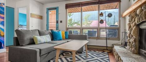 A cozy living room with a gray sofa, a wooden coffee table, and a patterned rug. There's a stone fireplace and a large window with a view of the outside.