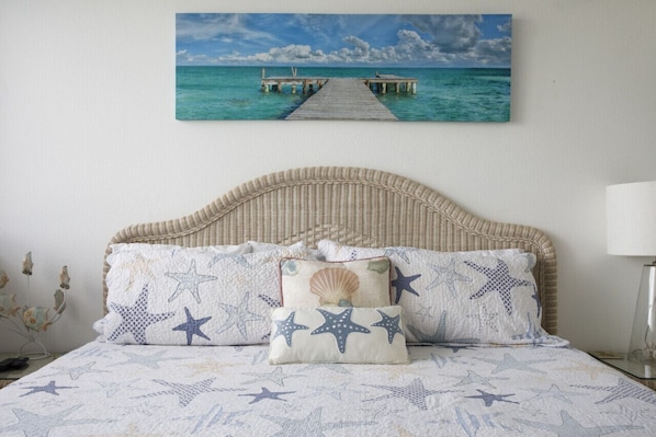 A bedroom with a bed featuring a starfish and seashell themed duvet cover, a wicker headboard, and a painting of a pier over turquoise water on the wall.