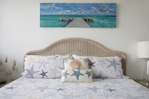 A bedroom with a bed featuring a starfish and seashell themed duvet cover, a wicker headboard, and a painting of a pier over turquoise water on the wall. - Gorgeous Moon Bay Condo Heated Pool Beach (Key Largo)