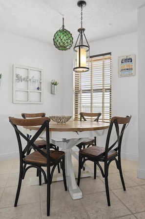 A dining area with a round table, four wooden chairs, a hanging green light fixture, and a window with blinds. - Gorgeous Moon Bay Condo Heated Pool Beach (Key Largo)