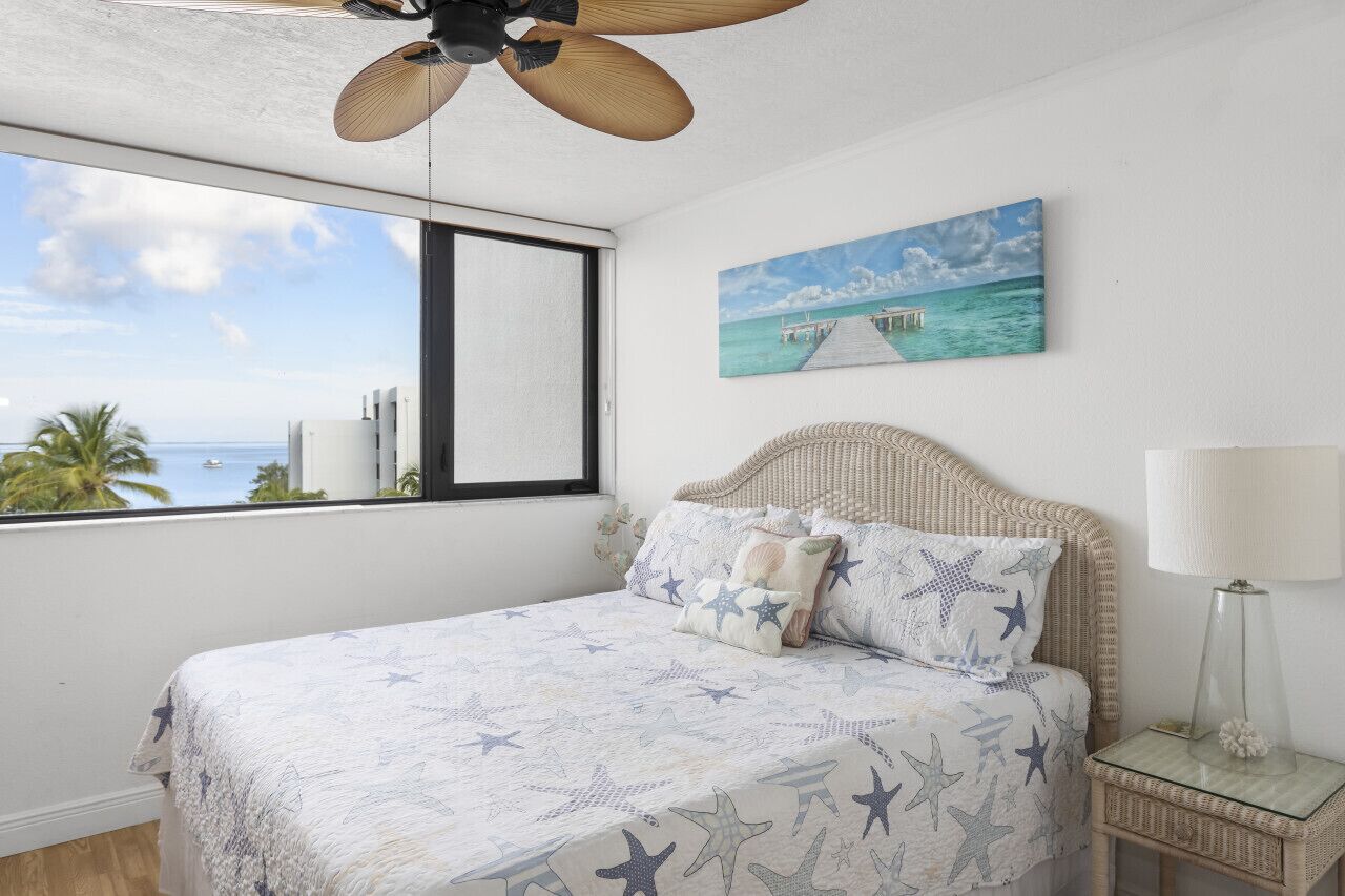 Une chambre à coucher avec un lit, un ventilateur de plafond, une fenêtre donnant sur l’océan et un tableau accroché au mur.