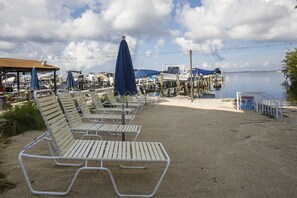 A beachside area with metal lounge chairs and umbrellas, a sandy shore, and a marina in the background.