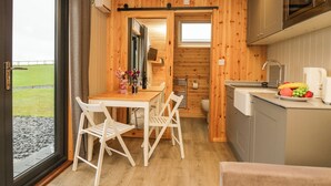 Cocina compacta con paredes de madera, zona de comedor con sillas blancas y vista a un campo de césped a través de una puerta de cristal.