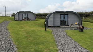 Dos modernas cabañas de techo gris y forma semicircular, con bancos de madera y mesas de picnic, ubicadas en un césped con un sendero de grava.