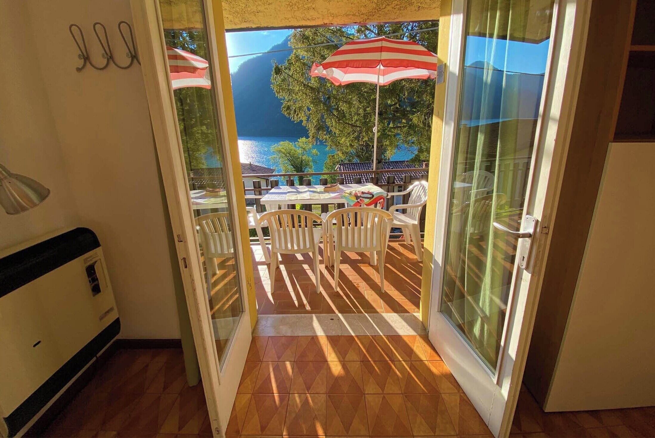 An open sliding glass door leading to an outdoor patio with a table and chairs, a red and white striped umbrella, and a view of a lake and mountains.
