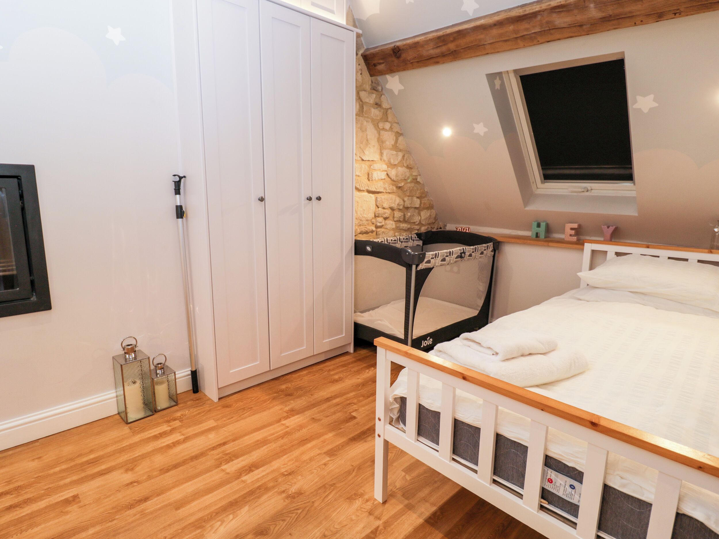 A bedroom with a wooden floor, a white bed, a stone wall, a window, and a wardrobe.
