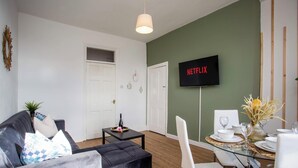 Una sala de estar con sofá, una mesa de centro, una mesa de comedor para dos personas y un televisor montado en la pared.