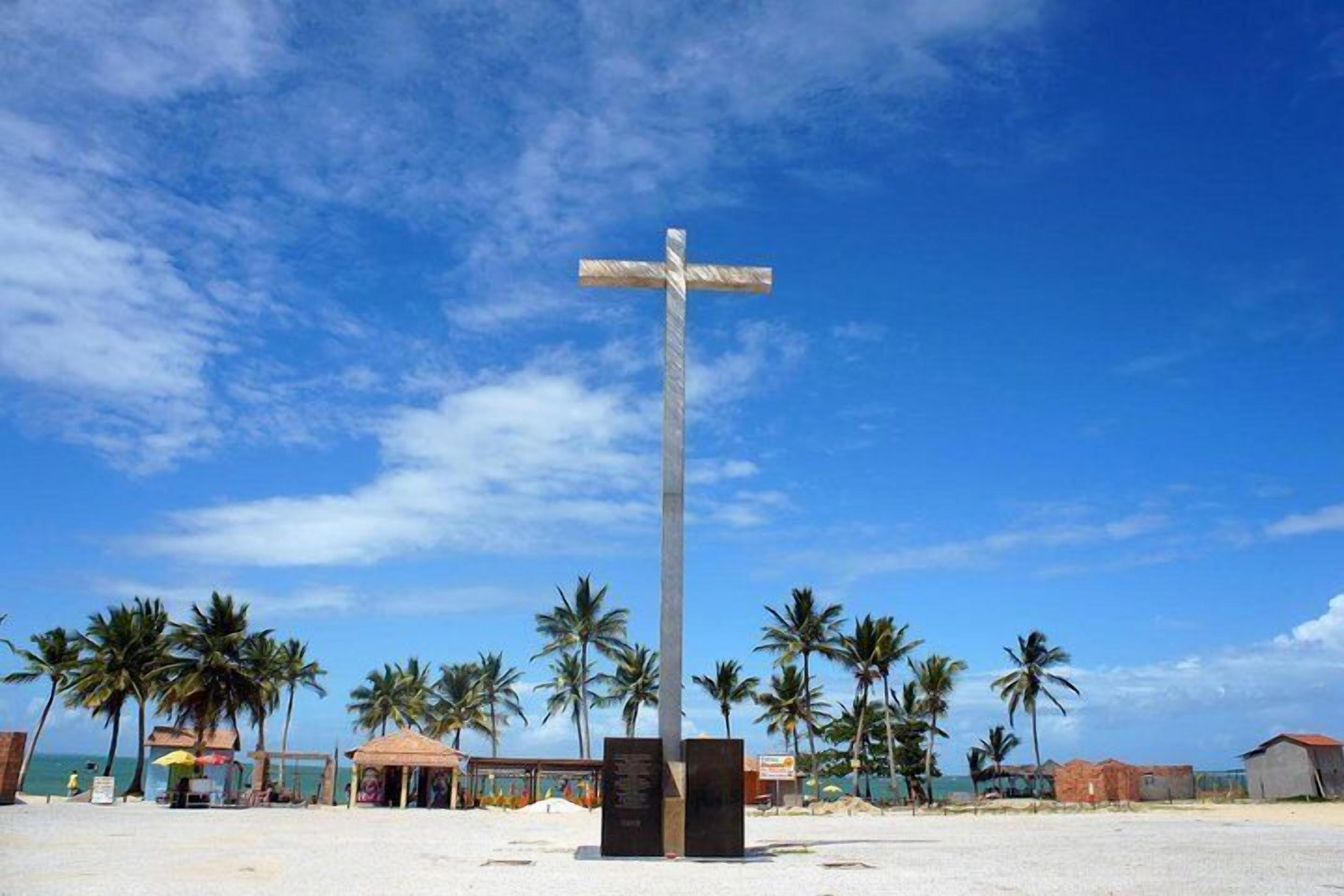 A tall cross monument on a beach with palm trees and buildings in the background.