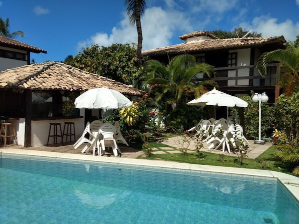 Zona de piscina con sillas reclinables blancas y sombrillas, rodeada de exuberante vegetación y un edificio con tejado de tejas.
