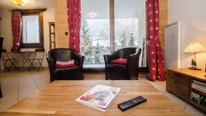 A room with two black chairs, a wooden table with a magazine and remote control, a window with red curtains, and a radiator on the right. - Flat with parking and balcony in Centre (Chamonix-Mont-Blanc)