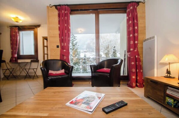 Une pièce avec deux chaises noires, une table en bois sur laquelle se trouvent un magazine et une télécommande, une fenêtre avec des rideaux rouges et un radiateur sur le côté droit.