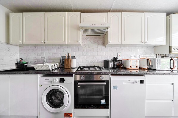 A compact kitchen with white cabinets, a built-in oven, a washing machine, and a microwave. - Cozy London Apartment for -3 (London)