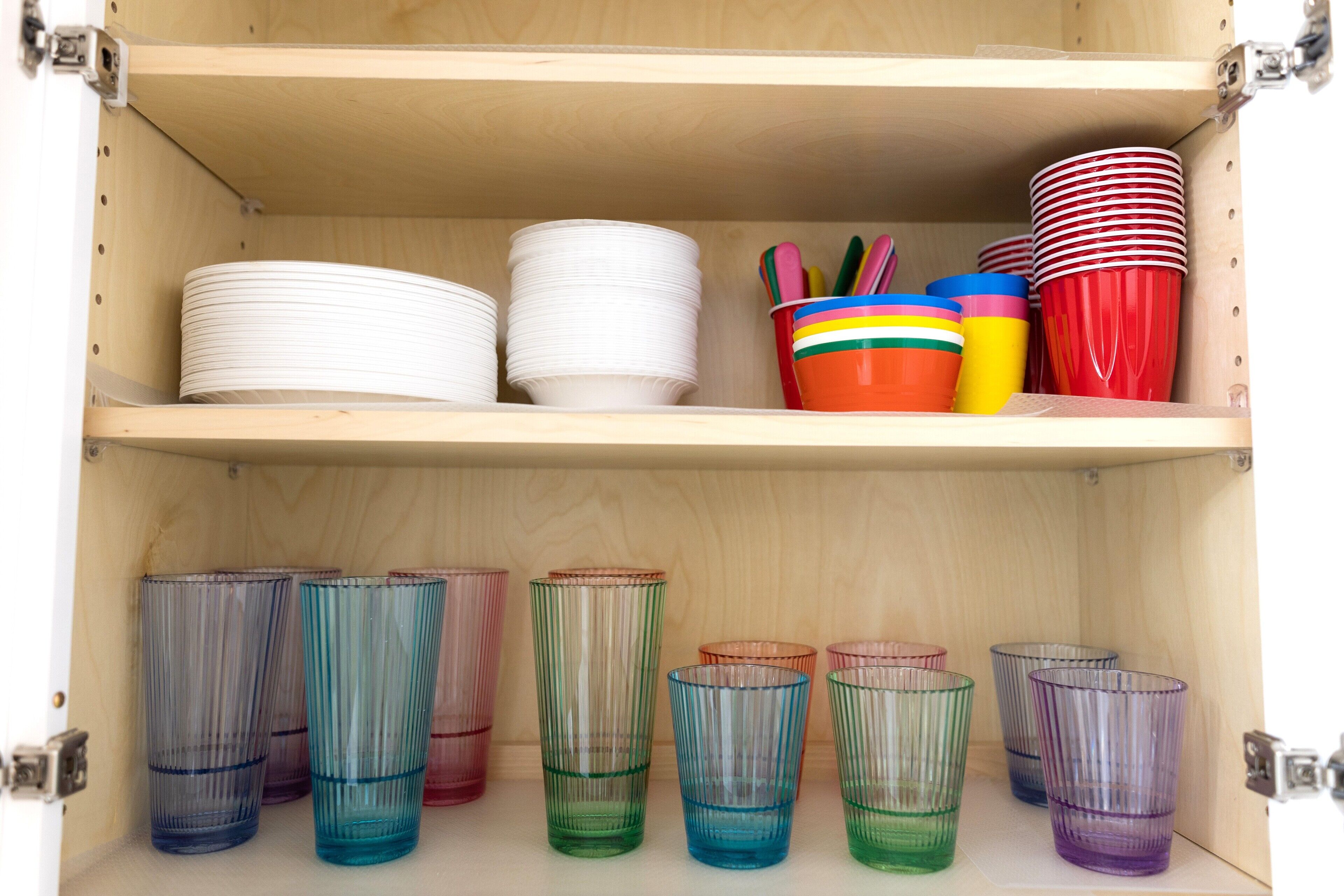 A wooden cabinet with shelves holding white plates, colorful cups, and transparent glasses.