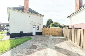 Ein Wohnhaus mit gepflasterter Terrasse, Holzzaun und einem weißen Gebäude mit schwarzem Sockel.