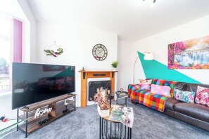 Een moderne woonkamer met een grote flatscreen-tv, een open haard, een kleurrijke bank en een schilderij aan de muur.
