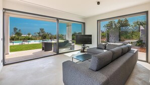 Uma sala de estar moderna com um sofá cinzento, uma televisão de tela plana e uma porta de vidro que dá para uma área externa com piscina.