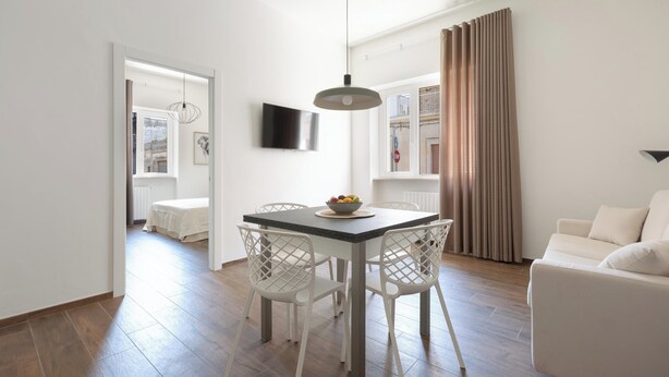 A modern living room with a dining area, white furniture, a flat-screen TV, and a large window with curtains.