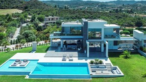 Una casa moderna de dos pisos con una amplia piscina, mobiliario de exterior y vistas del paisaje circundante.