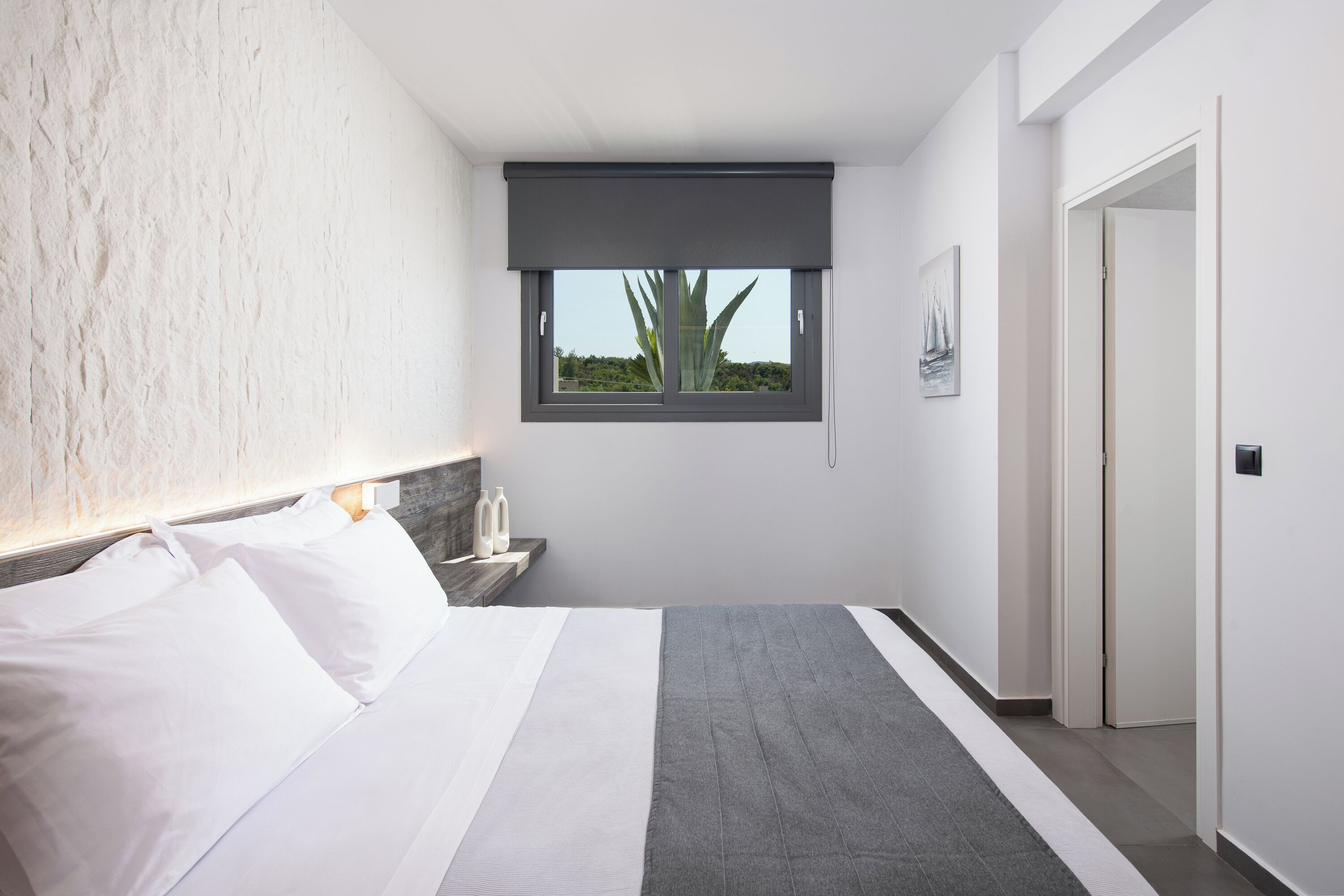 Una habitación de hotel moderna con una cama grande, una mesita de noche, un ventanal con vistas y una puerta.