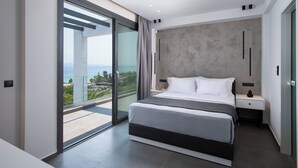 A modern bedroom with a large bed, a nightstand, and a view of the sea through a sliding glass door.