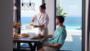 A breakfast setting with a view of the ocean, a dining table with plates of food, and a person serving orange juice.