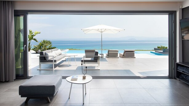 A modern living room with a large glass door leading to a pool area with outdoor furniture and a view of the sea.