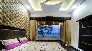 A modern bedroom with a geometric patterned wall, a large bed with a patterned bedspread, a ceiling fan, and a view of the outdoors through a window. - Raj Sea Front Beach Resort 102 (Mangaluru)