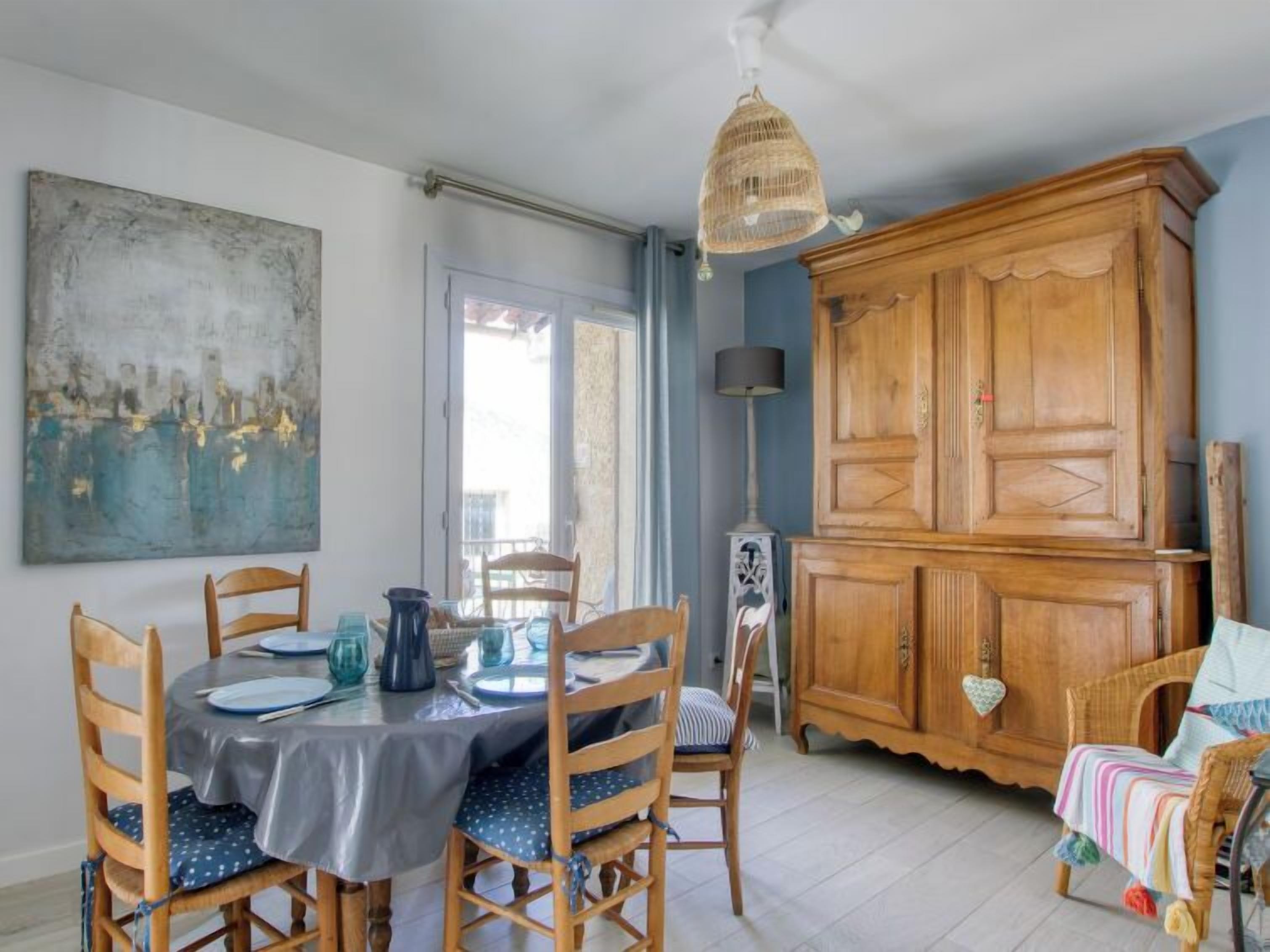 A dining area with a round table set for four, surrounded by wooden chairs. A large wooden cabinet is against the wall, and a painting is hung above the table.