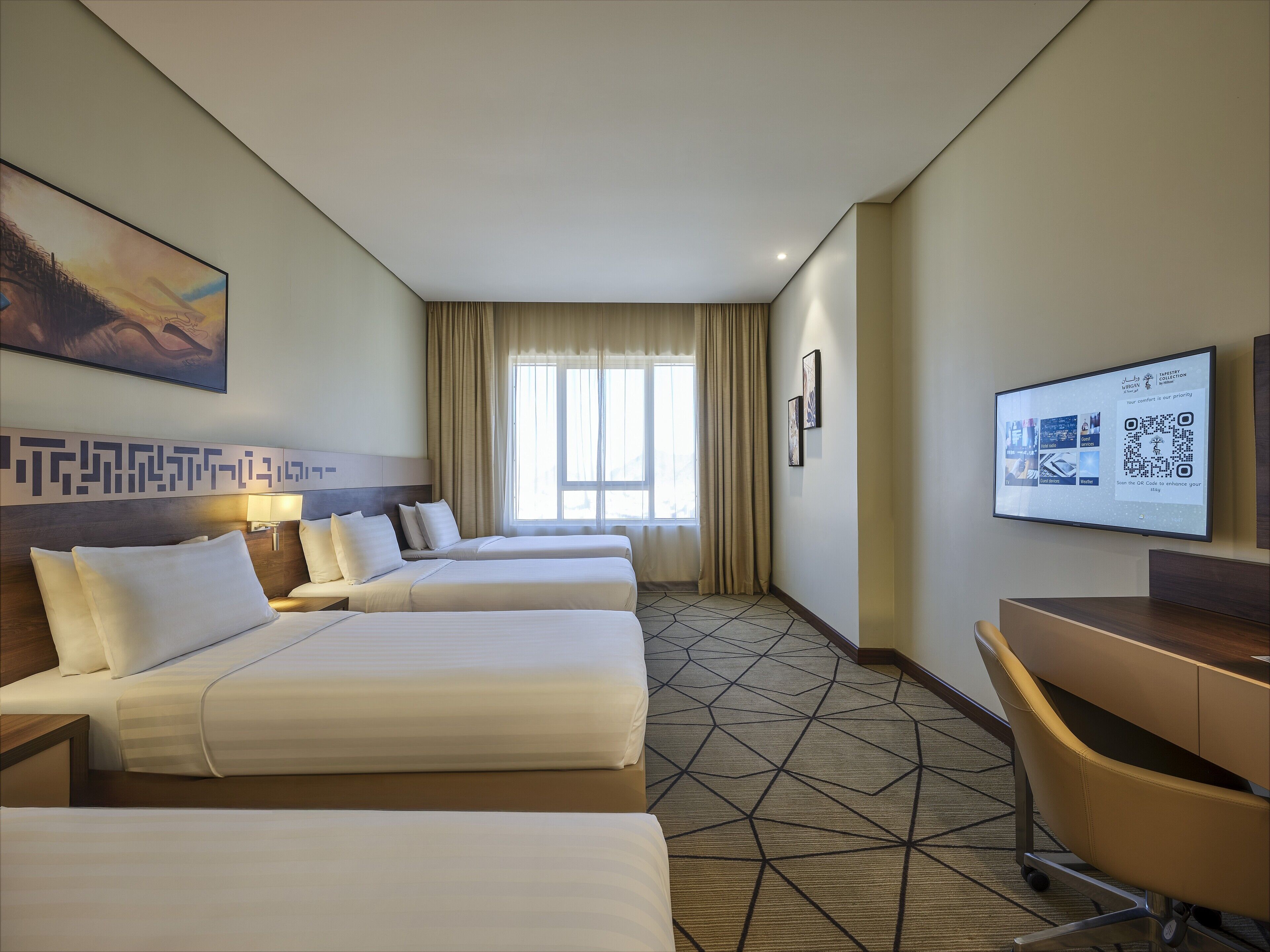 Une chambre d’hôtel avec deux lits, un bureau, une chaise, une grande fenêtre avec des rideaux, un tableau au mur et un écran numérique affichant des informations.